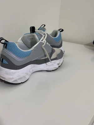 Chunky grå och blå sneakers - Säljer ett par chunky sneakers i grått, vitt och ljusblått med meshdetaljer och tjock sula. Skorna har vita snören och svarta dragflikar bak. Perfekta för dig som gillar sportig och trendig stil.