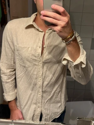 Beige linneskjorta  - Säljer en beige skjorta i linne från H&M med klassisk krage och bröstficka. Skjortan har långa ärmar och en avslappnad passform, perfekt för en chill och stilren look. Materialet är lätt och andas bra.