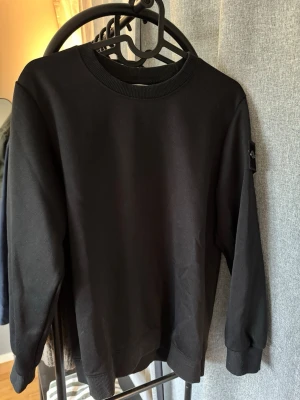 Svart sweatshirt från Stone Island - Säljer en svart sweatshirt från Stone Island med den klassiska loggan på vänster ärm. Tröjan har rund halsringning, ribbade muddar vid ärmslut och nederkant samt är gjord i mjukt bomullsmaterial. Perfekt för en clean och stilren look.