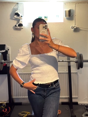 Vit smockad blus med puffärm - Supersöt vit blus med smockad kropp och puffiga korta ärmar. Fyrkantig ringning och lätt volangkant nedtill ger en trendig och feminin look. Perfekt till jeans eller kjol för en fräsch stil. Drar in midjan! 