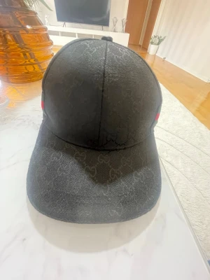 Svart keps från Gucci med GG-mönster - Säljer en svart keps från Gucci med det klassiska GG-monogrammet över hela kepsen. På sidan finns en grön och röd rand i tyg, typiskt för Gucci. Kepsen har böjd skärm och justerbar baksida. Perfekt för dig som vill ha en stilren och lyxig accessoar.
