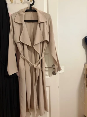 Beige trenchcoat med knytskärp - Stilren beige trenchcoat med bred krage och knytskärp i midjan. Långa ärmar och avslappnad passform ger en klassisk look. Perfekt för lager på lager och enkel att matcha med olika outfits. Passar strl 36/38