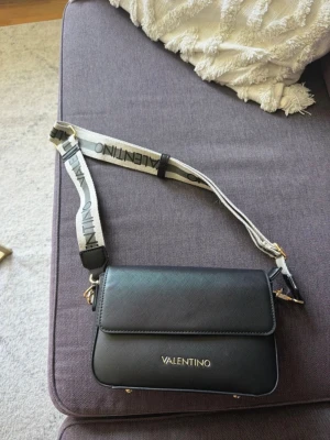 Svart axelväska från Valentino - Snygg svart axelväska från Valentino med justerbar bred axelrem i vitt och svart med logotyp. Väskan har stilren design, guldfärgade detaljer och är tillverkad i ett slätt konstläder. Perfekt för dig som vill ha en trendig och praktisk väska.
