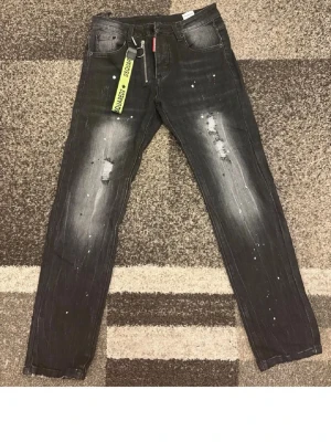 Svarta Dsquared2 jeans med detaljer - Säljer ett par svarta jeans från Dsquared2 med slitningar, vita färgstänk och en cool gul nyckelring. Jeansen har raka ben, klassisk femficksmodell och en snygg tvättad look. Perfekta för dig som gillar streetwear och vill sticka ut.