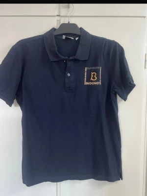Mörkblå pikétröja från Burberry - Klassisk mörkblå pikétröja från Burberry med gul broderad logga och text på bröstet. Tröjan har krage, korta ärmar och knappar framtill. Perfekt för en stilren och avslappnad look.
