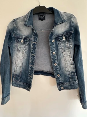 Blå tvättad jeansjacka från SALT - Snygg blå jeansjacka från Vally med tvättad look, stretchig denim och klassisk krage. Jackan har två bröstfickor med lock och knappar, samt silverfärgade knappar framtill. Långärmad modell i mjukt jeansmaterial, perfekt för en avslappnad stil. Jackan är ny/oanvänd -SAKNAR TAGS. Inköpt på Pondus för ca 1000kr.