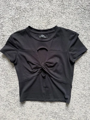 Svart crop top med twist framtill - Snygg svart crop top med korta ärmar och twistad detalj framtill samt en liten cut-out ovanför tvisten. Tillverkad i mjukt bomullsliknande material och har en tight passform. Perfekt till jeans eller kjol för en trendig look.