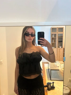 Svart volangig axelbandslös topp - Säljer en svart axelbandslös topp med volangdetaljer och luftig passform. Säljer den då den inte kommer till användning, och är använd fåtalet gånger! Toppen är croppad och har en bandeau-stil💗