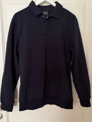 Mörkblå långärmad pikétröja från H&M premium  - Stilren mörkblå långärmad pikétröja från H&M premium med klassisk krage och tre knappar framtill. Tröjan har ribbade muddar vid ärmslut och är gjord i mjukt bomullstyg. Perfekt för en avslappnad men snygg look.