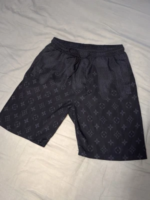 Mörkblå shorts med monogrammönster - Snygga mörkblå shorts med klassiskt monogrammönster och elastisk midja med snörning. Shortsen har en loose passform och är perfekta för sommaren. Materialet känns lätt och bekvämt, vilket gör dem sköna att bära hela dagen.