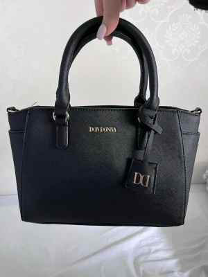 Svart handväska från Don Donna - Stilren svart handväska från Don Donna med två handtag och guldfärgade detaljer. Väskan har en klassisk, rektangulär form och är tillverkad i ett tåligt konstlädermaterial. Invändigt finns dragkedjefack och flera mindre fack för organisering. Perfekt för dig som gillar enkel och snygg design. Handväskan är aldrig använd och inga synliga tecken på användning❤️(Nypris 500kr)