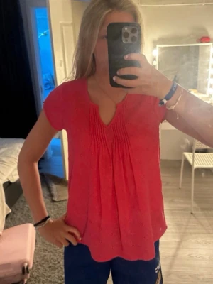 Rosa topp med broderade detaljer - Snygg rosa topp med korta ärmar och broderade detaljer. Toppen har en v-ringad hals med veckade partier framtill som ger en avslappnad och luftig känsla. Perfekt för en trendig och bekväm stil.