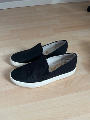 Svarta slip-on sneakers - Svarta slip-on sneakers från Sam Edelmam vit sula. Skorna har en enkel och stilren design utan snörning, vilket gör dem lätta att ta på och av. Ovandelen är i textil och sulan är i gummi. 