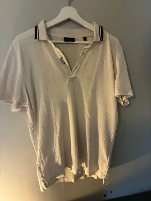 Vit piké från Gant med ränder - Vit pikétröja från Gant med diskreta diagonala ränder och klassisk krage med röd och marinblå rand. Kortärmad modell i mjuk bomull, perfekt för en clean och sportig stil. Knappslå framtill och relaxed fit. Hör av er vid frågor🔥