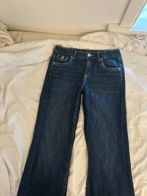 Mörkblå bootcut jeans med kontrastsömmar - Snygga mörkblå bootcut jeans med vita kontrastsömmar och klassiska fem fickor. Jeansen har knapp- och dragkedjestängning samt detaljerade bakfickor med lock. Perfekta för en avslappnad och trendig look.