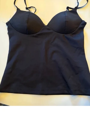 Svart body med tunna axelband - Snygg svart body med tunna justerbara axelband och vadderade kupor. Tillverkad i stretchigt material som sitter bekvämt mot kroppen. Perfekt att bära under kläder eller som ett statement-plagg. Enkel och stilren design utan synliga detaljer.
