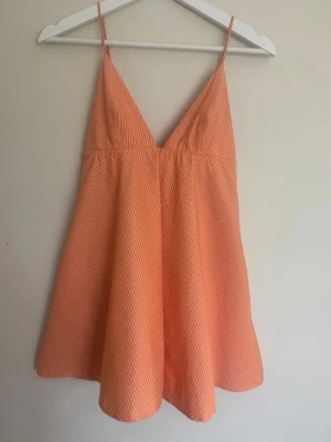 Randig orange kort sommarklänning - Söt kort klänning i orange med tunna vita ränder och smala axelband. Klänningen har en v-ringad front och ärmlös design, perfekt för varma dagar. Materialet känns lätt och luftigt, vilket gör den skön att bära under sommaren. Pris kan diskuteras