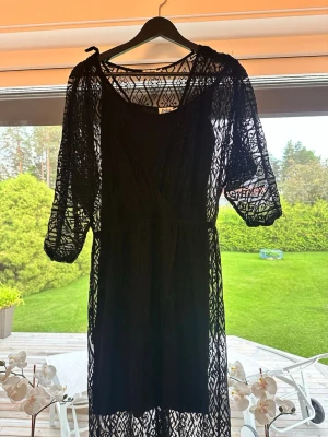 Svart omlottklänning i spets - Säljer en svart omlottklänning med halvtransparent spets och trekvartsärm. Klänningen har ett geometriskt mönster i spetsen och en v-ringad omlottdesign. Perfekt för dig som vill ha en unik och snygg look. Man kan även använda enbart spetsen som en cover up på stranden.
