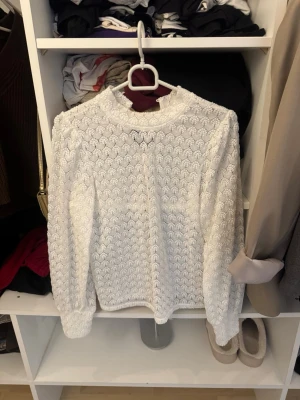Vit spetsblus med lång ärm - Säljer en vit spetsblus med långärmad design och hög krage. Blusen har ett vackert spetsmönster över hela plagget och en liten öppning vid halsen. Perfekt för dig som gillar romantisk och elegant stil.
