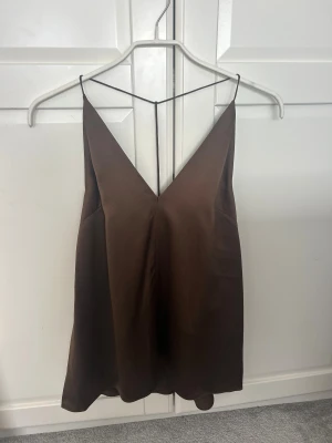 Dagmar silktopp  - Snygg brun silktopp från Dagmar. Toppen är ärmlös och axelbandslös med tunna band och djup v-ringning både fram och bak. Perfekt för dig som gillar minimalistisk och stilren design. Helt oanvänd med lapp.