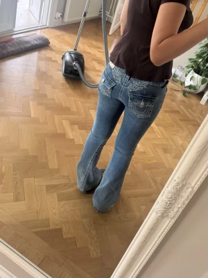 Blå bootcut jeans från True Religion - Säljer ett par blå bootcut jeans från True Religion med ikoniska kontrastsömmar och detaljerade bakfickor med lock och knappar. Jeansen har låg midja och klassisk femficksdesign. Perfekta för dig som gillar en avslappnad men trendig look.