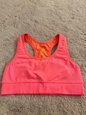 Neonrosa sport-bh  - Snygg neonrosa sport-bh från Esprit med brottarrygg och elastisk kant nedtill. Tillverkad i ett stretchigt och snabbtorkande material som passar perfekt till träning. Logga i silver på framsidan. Insidan är orange för en cool kontrast. +OANVÄND! (Prislapp kvar)