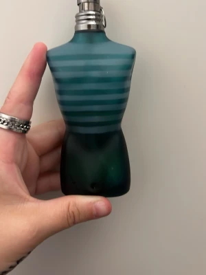 Jean Paul Gaultier Le Male parfym - Ikonisk parfymflaska från Jean Paul Gaultier formad som en manlig torso i blått glas med turkosa ränder. Flaskan har en metallkork och designen är riktigt unik och cool, perfekt för dig som gillar statement-detaljer på hyllan. Halva flaskan kvar