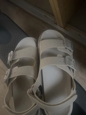 Vita/begie ish sandaler med dubbla spännen - Säljer ett par vita/begie aktiga sandaler med två breda remmar och justerbara spännen. Sandalerna har öppen tå och hälrem för extra stöd. Perfekta för sommaren och riktigt snygga på en vanlig sommar dag. ( Använd)