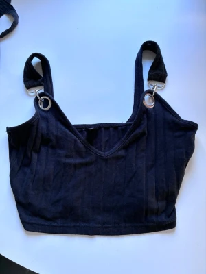 Svart ribbad croppad linnetopp - Snygg svart croppad linnetopp med ribbad struktur och breda axelband som fästs med silvriga ringdetaljer. Toppen har en v-ringad front och är perfekt för en trendig och avslappnad stil.