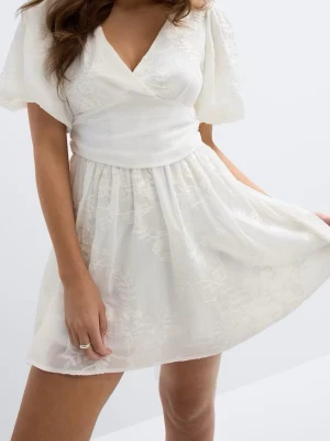 Vit omlottklänning med rosett - Superfin vit kortklänning med omlottliv, puffiga kortärmar och markerad midja. Klänningen har broderade blommor i tyget, v-ringning och en stor rosett bak. Tillverkad i ett lätt och luftigt material, perfekt för sommarens alla tillfällen.