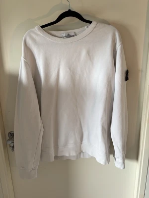 Vit sweatshirt från Stone Island - Säljer en clean vit sweatshirt från Stone Island med den klassiska svarta patchen på ärmen. Tröjan har rund halsringning och är tillverkad i mjukt bomullsmaterial. Perfekt för dig som gillar streetwear och enkel stil.