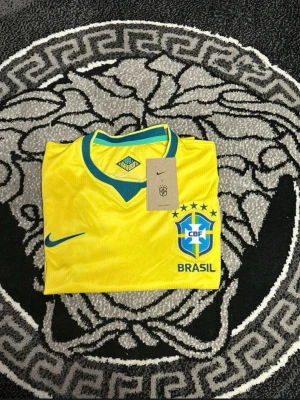 Gul Brasil Nike fotbollströja - Snygg gul Brasilien fotbollströja från Nike med blå detaljer och CBF-logga på bröstet. Tröjan har korta ärmar, rund halsringning med blå och grön kant samt Nike-logga på framsidan. Perfekt för dig som älskar fotboll och vill stötta Brasilien.
