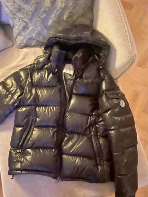 Moncler Maya  - Säljer en svart, glansig dunjacka från Moncler med huva och flera fickor. Jackan har en puffig design, dragkedja framtill och Moncler-logga på ärmen. Perfekt för kalla vinterdagar och riktigt snygg streetstil. Storlek 4 passar 3 alltså L perfekt 