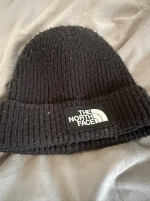 Svart ribbstickad mössa The North Face - Säljer en svart ribbstickad mössa från The North Face med uppvikt kant och stor logotyp framtill. Klassisk modell som passar perfekt till kalla dagar. Tillverkad i mjukt och värmande material.