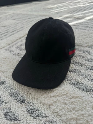 Svart keps från Gucci med röd-grön rand - Snygg svart keps från Gucci med klassisk böjd skärm och diskret GG-mönster i tyget. På sidan finns en ikonisk röd och grön rand. Justerbar rem baktill för perfekt passform. Perfekt accessoar för dig som gillar exklusiv streetwear.