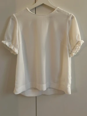 Vit blus med volangärm från Bondelid - Söt vit blus från Bondelid med puffiga korta ärmar och volangdetalj vid ärmslut. Blusen har en enkel, rak passform och rund halsringning. Perfekt att styla med jeans eller kjol för en fräsch look. En liten diskret Bondelid logga finns längst ner på ena sidan på blusen. Blusen är av 100% polyester och därför väldigt luftig och skön.