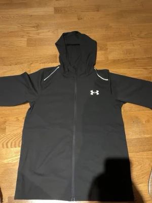 Svart Jacka  från Under Armour - Svart Jacka  från Under Armour med dragkedja framtill och vit logga på bröstet. Stora vita bokstäver med UNDER ARMOUR på ryggen. Jackan har huva och vita detaljer vid axlarna. Tillverkad i mjukt och stretchigt material, perfekt för en sportig look.
