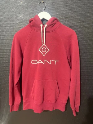 Röd hoodie från GANT - Snygg röd hoodie från GANT med stor vit logga och emblem på bröstet. Klassisk känguruficka framtill, ribbade muddar och vit dragsko i huvan. Tillverkad i mjukt bomullsmaterial som känns skönt mot huden. Perfekt för en avslappnad och trendig look.