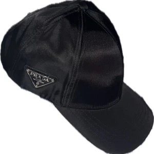 Svart keps från Prada i nylon - Snygg svart keps från Prada med klassisk böjd skärm och ikonisk metalltriangel-logga på sidan. Tillverkad i glansigt nylonmaterial som ger en exklusiv vibe. Perfekt för dig som vill ha en stilren och lyxig accessoar med streetkänsla.