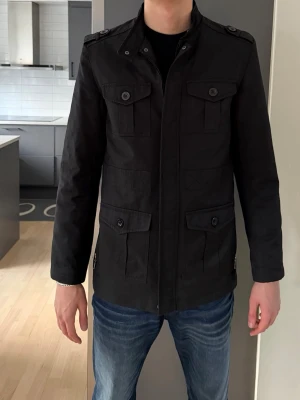 Overshirt - Stilren svart utilityjacka med fyra framfickor och knappar. Jackan har hög krage, axelklaffar och stängs med dragkedja och knappslå. Passformen är normal och materialet känns som bomull eller polyester. Perfekt till jeans och t-shirt.