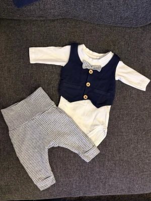 Bodyset med väst och byxor från Name It - Sött set från Name It med långärmad vit body, fastsydd marinblå väst och matchande fluga med ränder. Träknappar framtill ger en dressad look. Tillhörande byxor i vitt och blått randigt tyg. Perfekt för speciella tillfällen eller när du vill klä upp din lilla. Ny  köptes i december men hann inte använda den. Har endast tvättat den. 