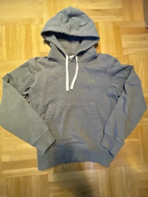 Grå Lacoste hoodie - Grå hoodie från Lacoste med klassisk krokodil-logga på bröstet. Tröjan har huva med vita dragsnören och en stor magficka. Perfekt för en avslappnad och stilren look. Passar ca 165. Rabatt vid snabb affär.