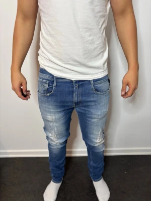 Blå slim fit jeans med slitningar Anbass - Snygga blå slim fit jeans med slitna detaljer och tvättade partier på låren och knäna. Klassisk femficksmodell med dragkedjegylf och smal passform. Perfekta för en avslappnad och trendig stil. Anbass