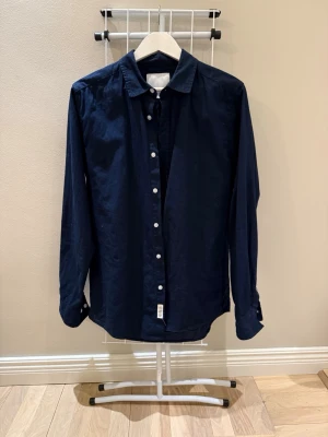 Mörkblå skjorta med knappar - Snygg mörkblå skjorta med klassisk krage och vita knappar framtill. Skjortan har lång ärm och är tillverkad i ett mjukt bomullstyg som känns skönt mot huden. Perfekt för dig som gillar en clean och stilren look.