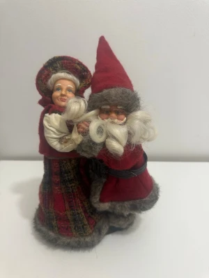 Tomtepar i tyg och ull - Charmigt tomtepar i klassisk julstil, tillverkade av tyg och ull. Tomten har röd luva, röd rock med grå pälskant och svart bälte, medan tomtemor bär röd och grön rutmönstrad klänning med huva. Båda har lockigt vitt hår och skägg.