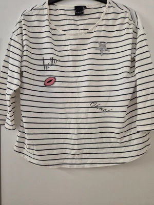 Tröja - Vit tröja med svarta horisontella ränder från Kappahl. Tröjan har broderade detaljer som en stjärna, ett par röda läppar och texten 'hello' och 'oh la la' på framsidan. Rund halsringning och trekvartsärm. Tillverkad i mjukt bomullsmaterial. Har några hål och lite gult underarmarna.