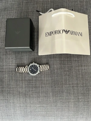 Emporio Armani silverklocka - Stilren klocka från Emporio Armani med silverfärgad länk i metall och svart urtavla. Klockan har kronograf-funktion, datumvisning och tydliga indexmarkeringar. Kommer med originalbox och Emporio Armani-påse. Perfekt accessoar för dig som gillar klassisk design. Den är justerbar, jag har delar om man vill göra den större eller mindre. 