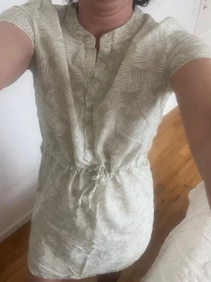 Ljusgrön kortklänning med knyt i midjan - Kortklänning ljusgrönt med vitt bladliknande mönster. Klänningen har korta ärmar, v-ringad hals med knappar och ett knytband i midjan för en snygg passform. Perfekt för varma dagar och enkel att styla.