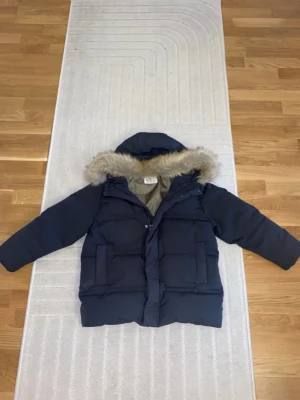 Mörkblå pufferjacka med fuskpäls - Säljer en mörkblå pufferjacka från Zara med avtagbar huva och fuskpäls. Jackan har två stora fickor framtill och en mjuk, olivgrön insida. Perfekt för kalla vinterdagar och riktigt snygg att bära utomhus.