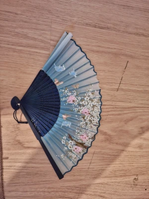 Blå solfjäder med blommönster - Snygg blå solfjäder med handtag i mörkblå plast och tyg i olika blå nyanser. Dekorerad med vita och rosa blommor samt fjärilar och små fåglar. Perfekt accessoar för att ge din stil en unik touch.